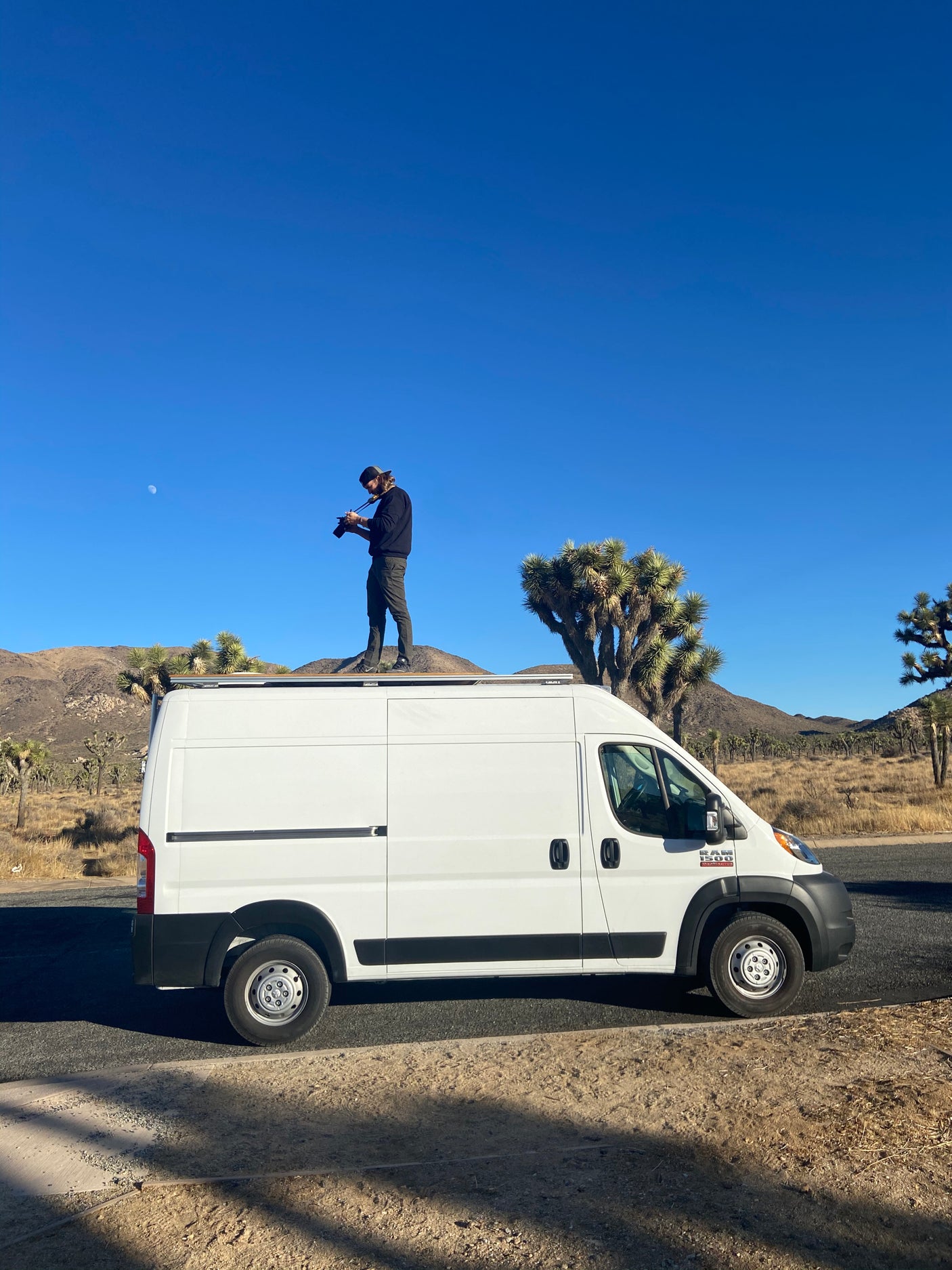 Man on RV rental campervan roof, photographing Joshua Tree desert scenery with visible moon.