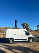 Man on RV rental campervan roof, photographing Joshua Tree desert scenery with visible moon.