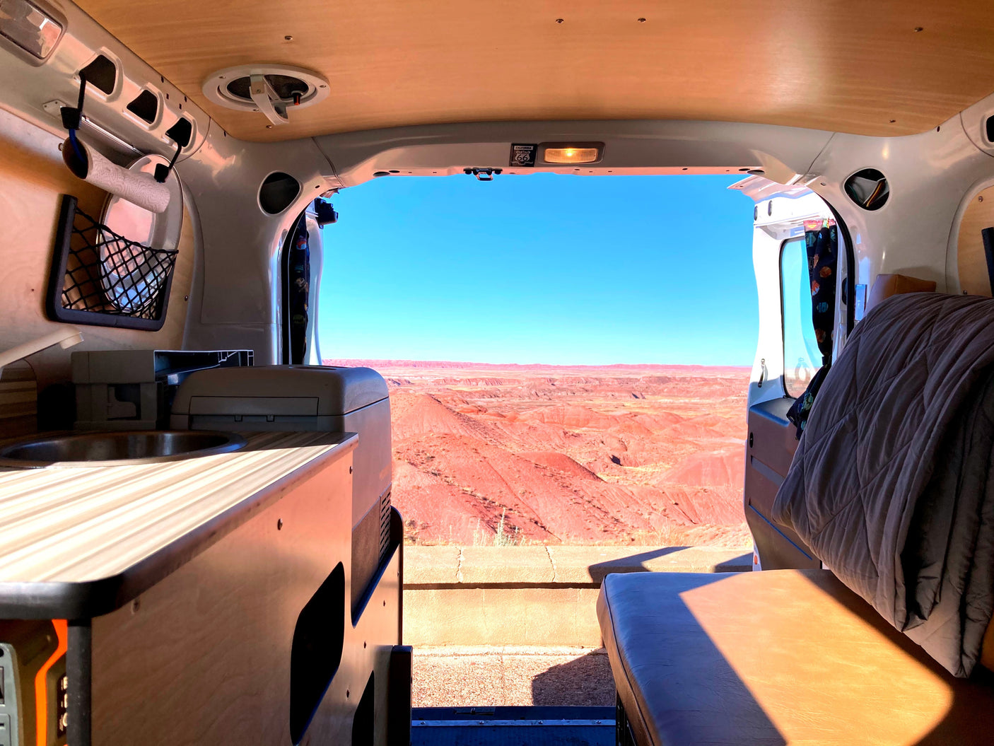 RV rental interior: Sink, bed, and panoramic view of red desert hills under a clear blue sky from a camper van.