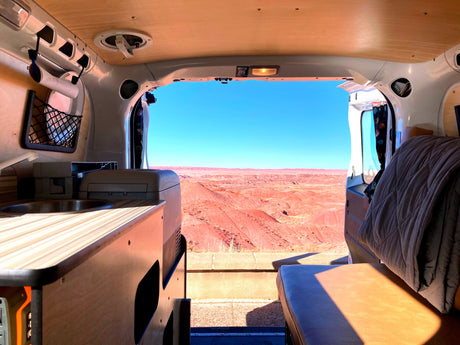 RV rental interior: Sink, bed, and panoramic view of red desert hills under a clear blue sky from a camper van.