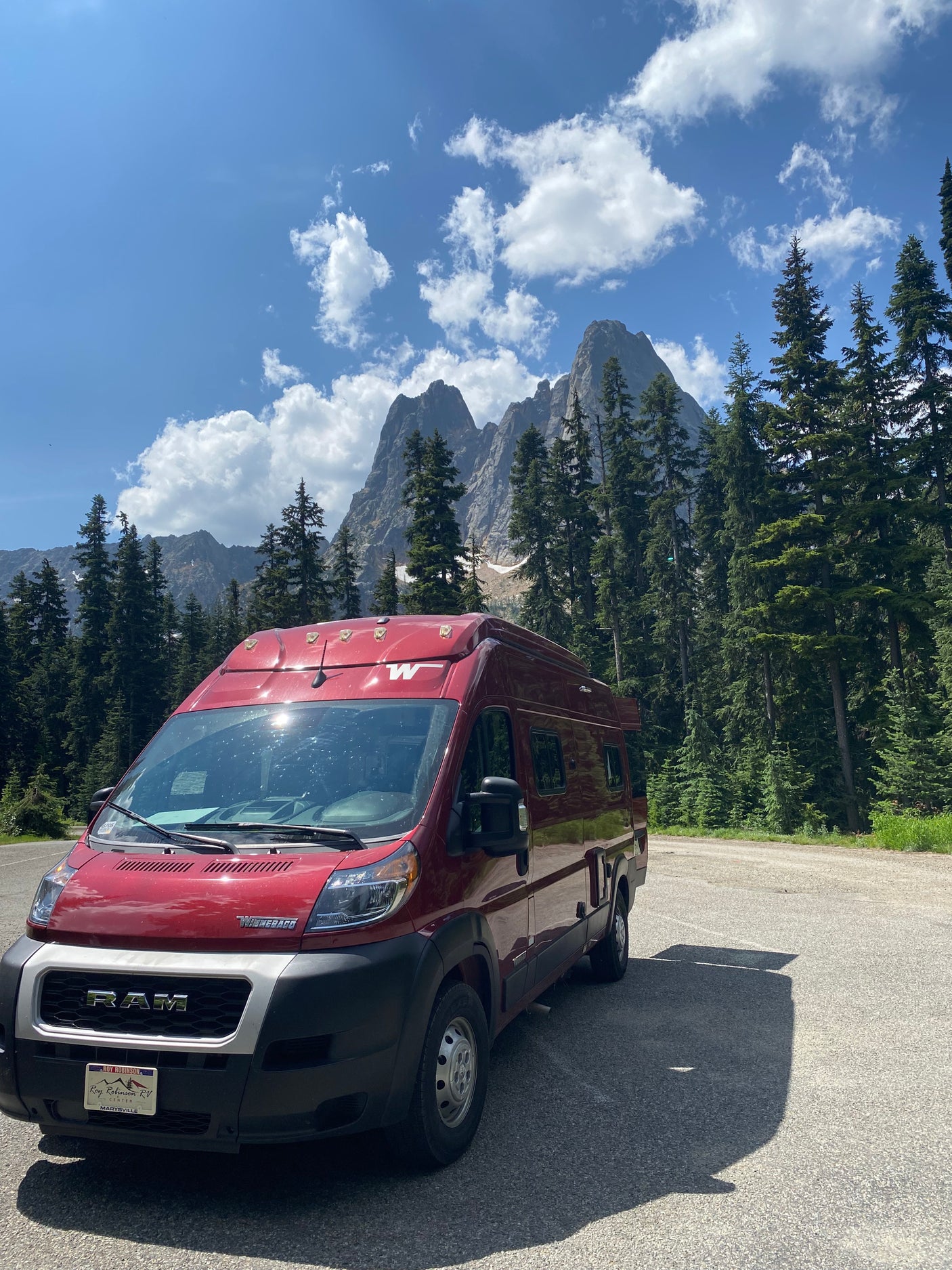 A red Winnebago rv rental van is parked on asphalt in a scenic mountain area with tall evergreen trees under a blue, cloudy sky.