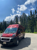 A red Winnebago rv rental van is parked on asphalt in a scenic mountain area with tall evergreen trees under a blue, cloudy sky.