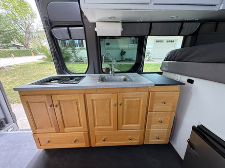 This RV rental interior features a functional kitchen with a sink, two-burner stovetop, and wood cabinets, plus a bed area, designed for compact travel.