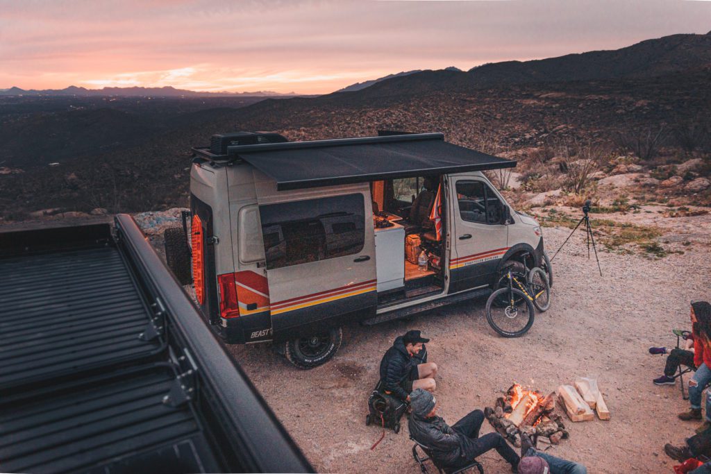  on a desert trail. Roof rack & ladder visible.|Adventure-ready RV rental! This off-road camper van with bright roof lights is perfect for exploring at sunset.|Front view: A tan & black Mercedes Sprinter adventure van — photo 4
