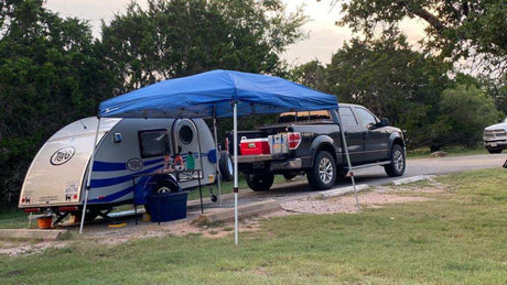 RV rental: A T@G travel trailer and pickup truck are parked at a campsite with a blue canopy, ready for outdoor adventures.