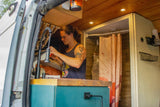 Inside a custom RV rental, a man uses the sink. Features compact kitchen, shower, and smart storage.