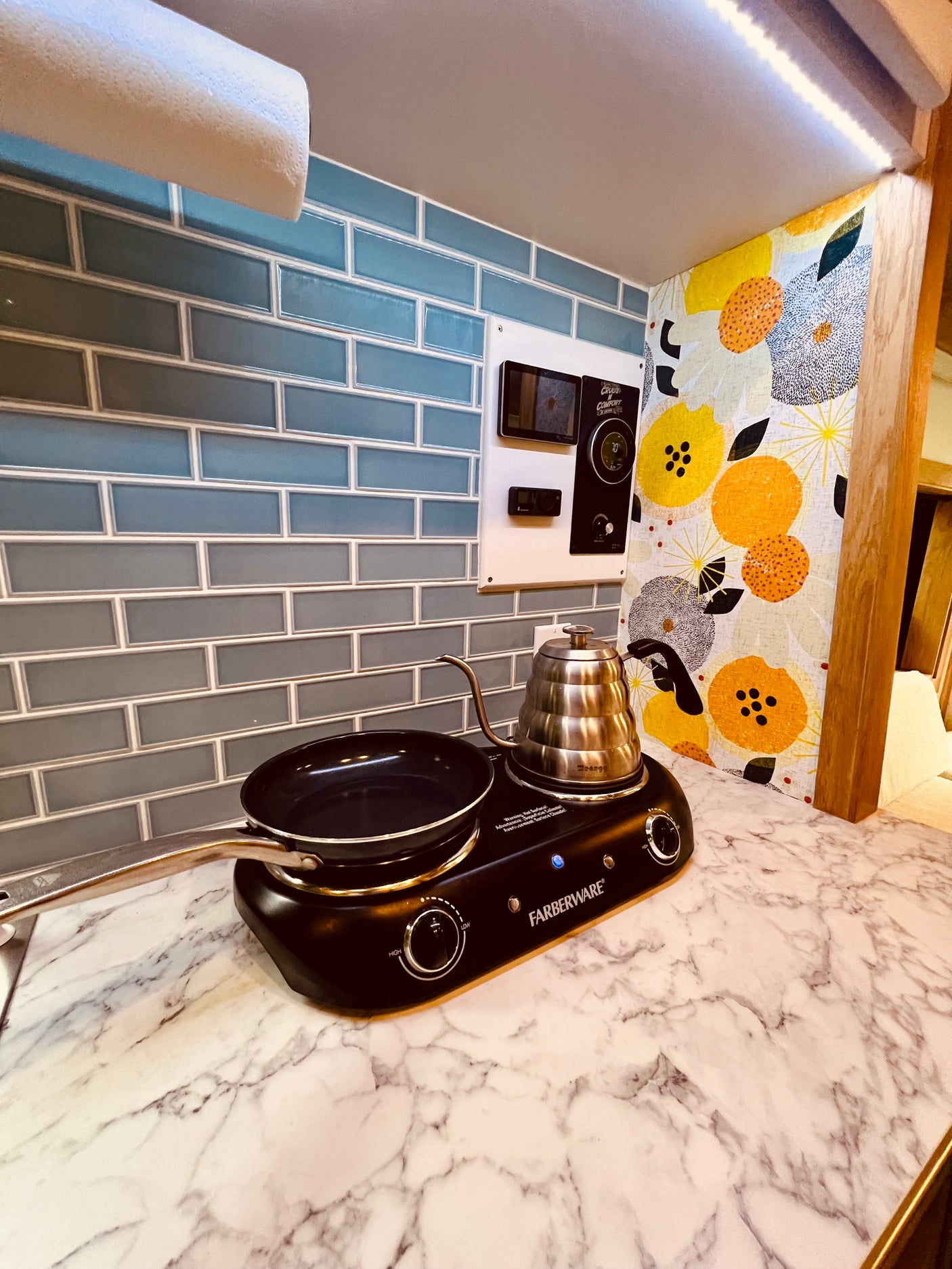RV rental kitchen: Compact setup with Farberware cooktop, pan, kettle, blue tile backsplash, and control panel.