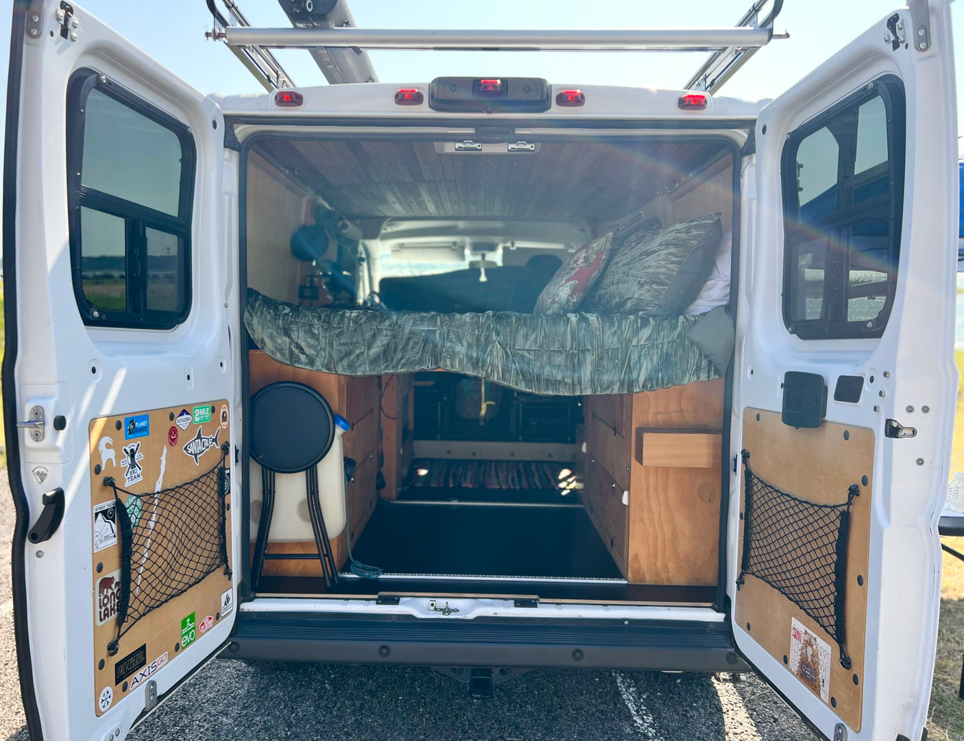 Open rear doors of an rv rental reveal a cozy camper van interior with a raised bed, wooden paneling, and accessible storage below.