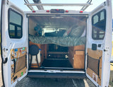 Open rear doors of an rv rental reveal a cozy camper van interior with a raised bed, wooden paneling, and accessible storage below.
