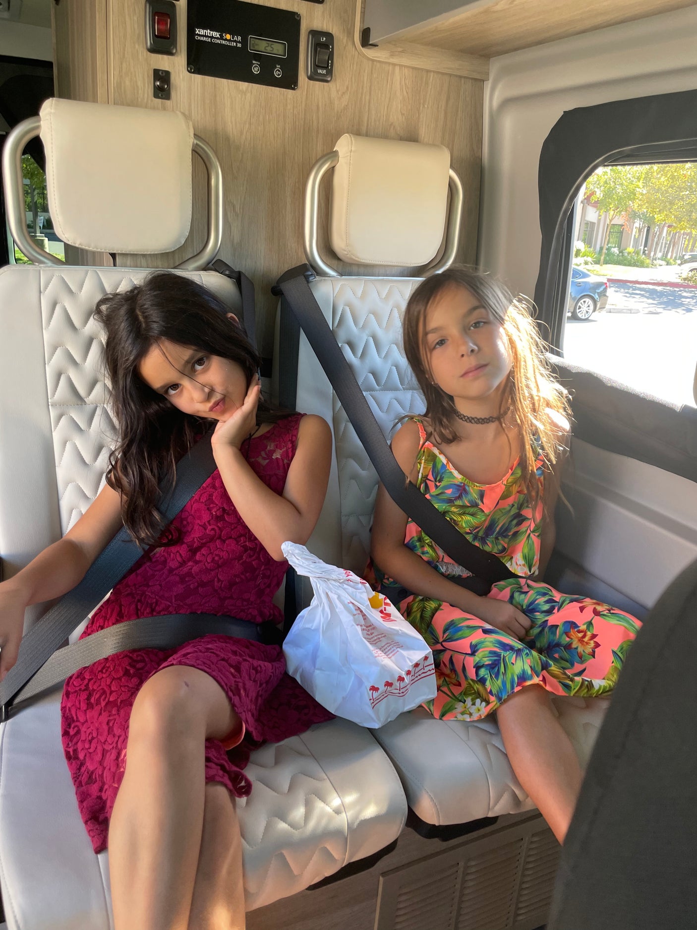 Two girls buckled in patterned seats of an RV rental, with an In-N-Out bag. A solar charge controller is visible.
