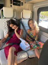Two girls buckled in patterned seats of an RV rental, with an In-N-Out bag. A solar charge controller is visible.