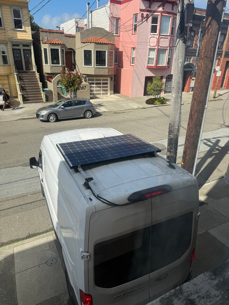 An RV rental van with a rooftop solar panel is parked on a sunlit city street, with houses in the background.