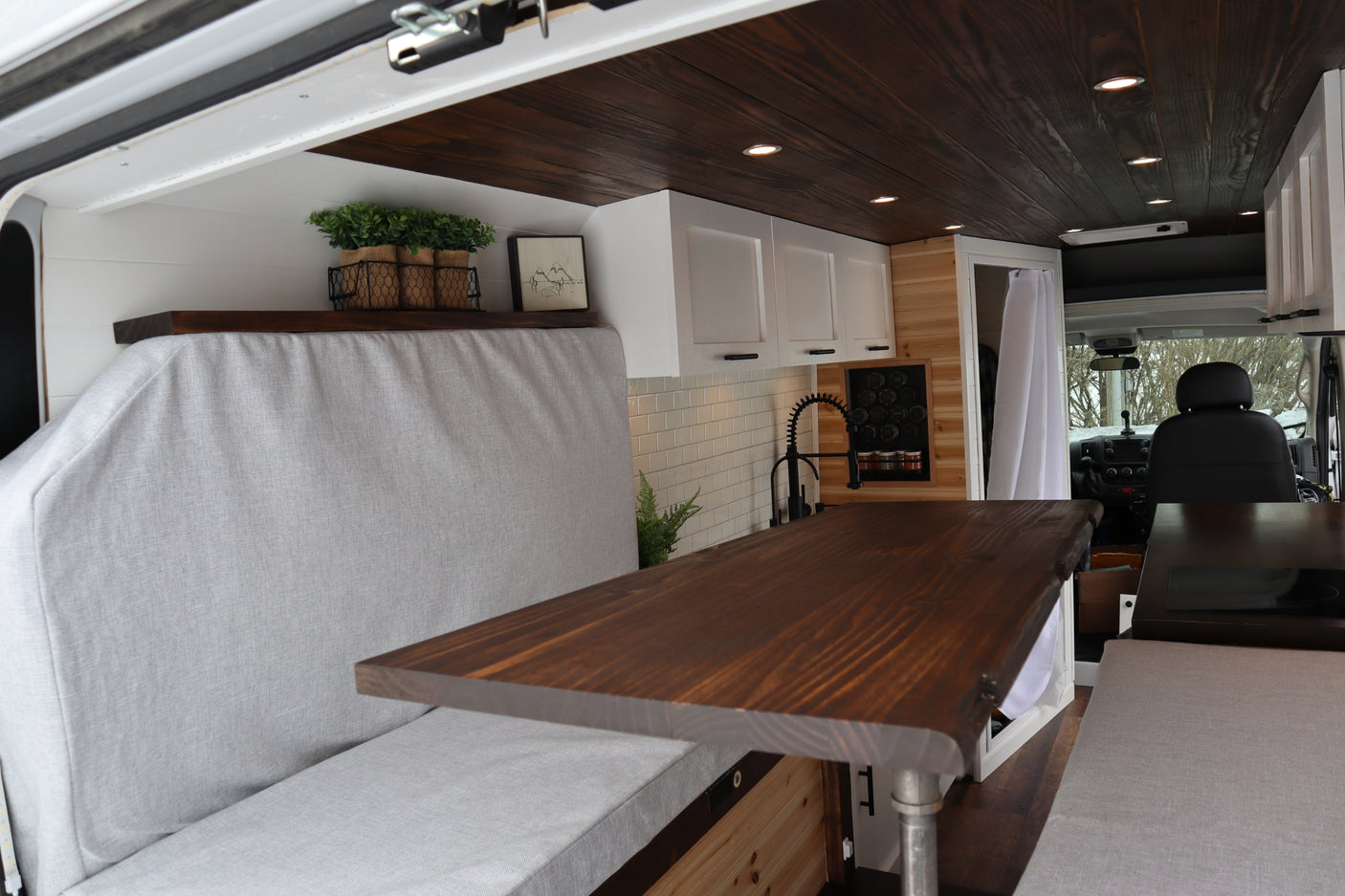 Experience a custom RV rental! This cozy interior features a warm wooden ceiling, white cabinets, and comfy grey seating with a dining table.