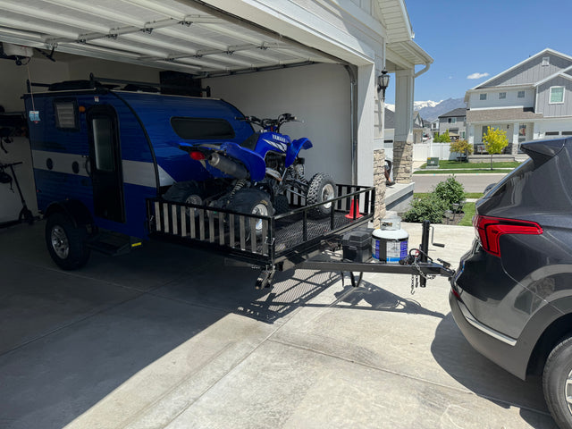 Blue teardrop RV, ATV on trailer hitched to SUV in a driveway. Perfect setup for your next RV rental adventure.