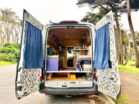 RV rental campervan with open rear doors showing its cozy interior setup amidst trees.