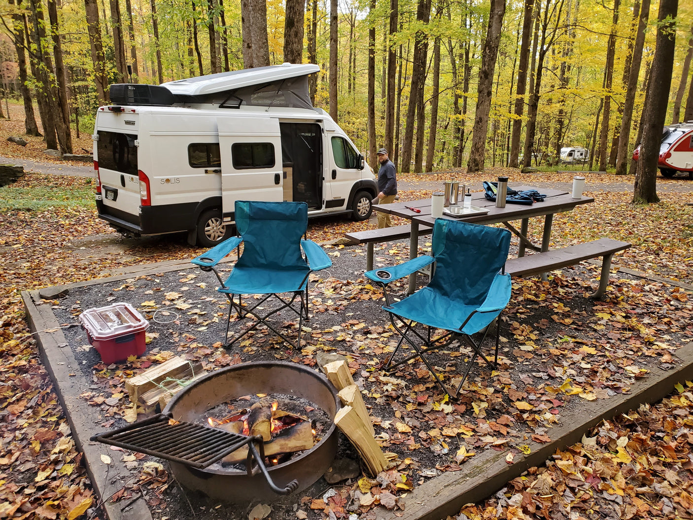 **RV rental** camper van at a cozy autumn campsite with a roaring campfire, picnic table, and vibrant fall foliage.