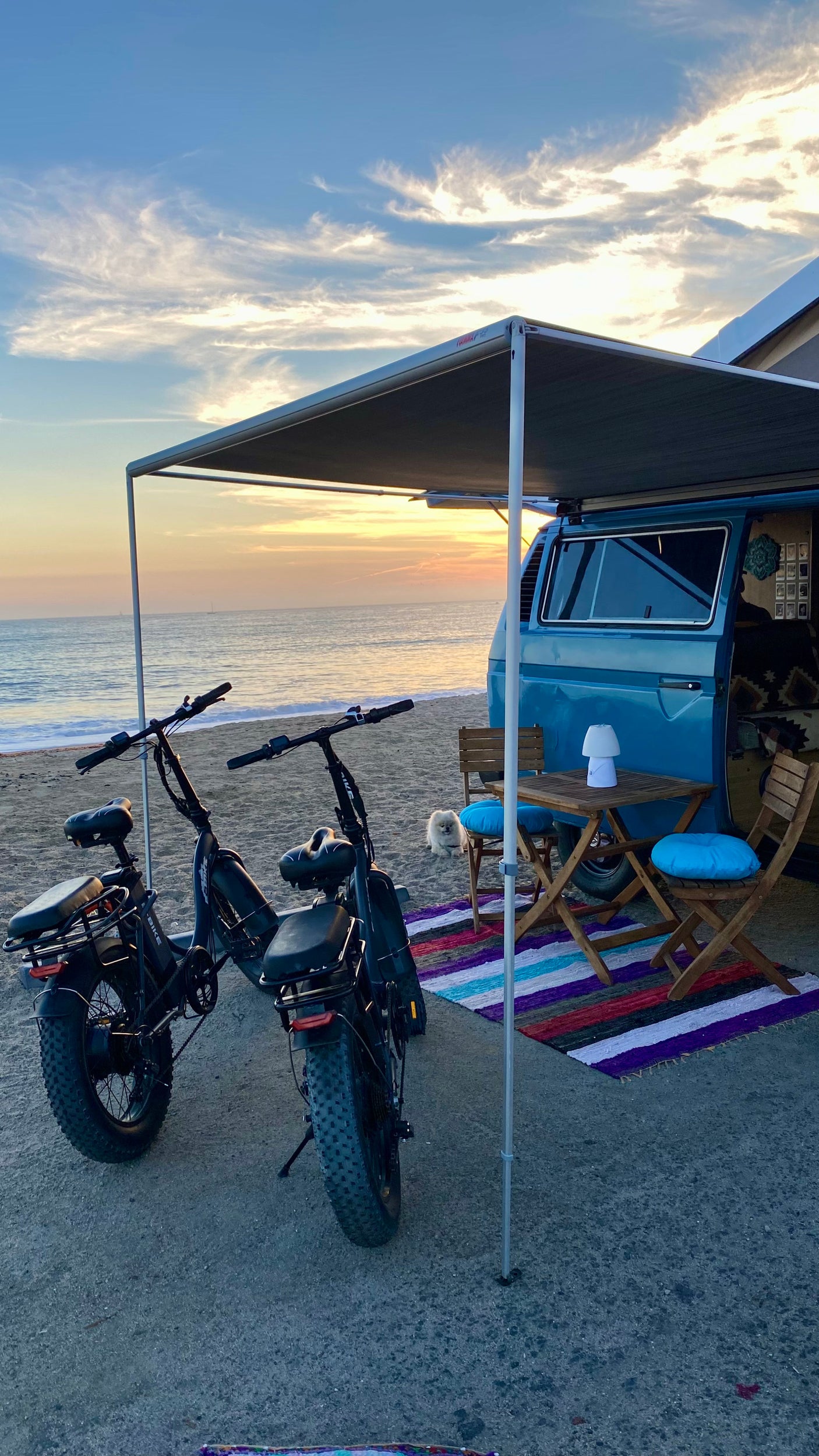 Sunset bliss: Blue camper van RV rental, two fat-tire e-bikes & beach setup with table, chairs, rug. Small dog joins the view!