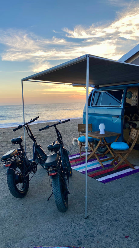 Sunset bliss: Blue camper van RV rental, two fat-tire e-bikes & beach setup with table, chairs, rug. Small dog joins the view!