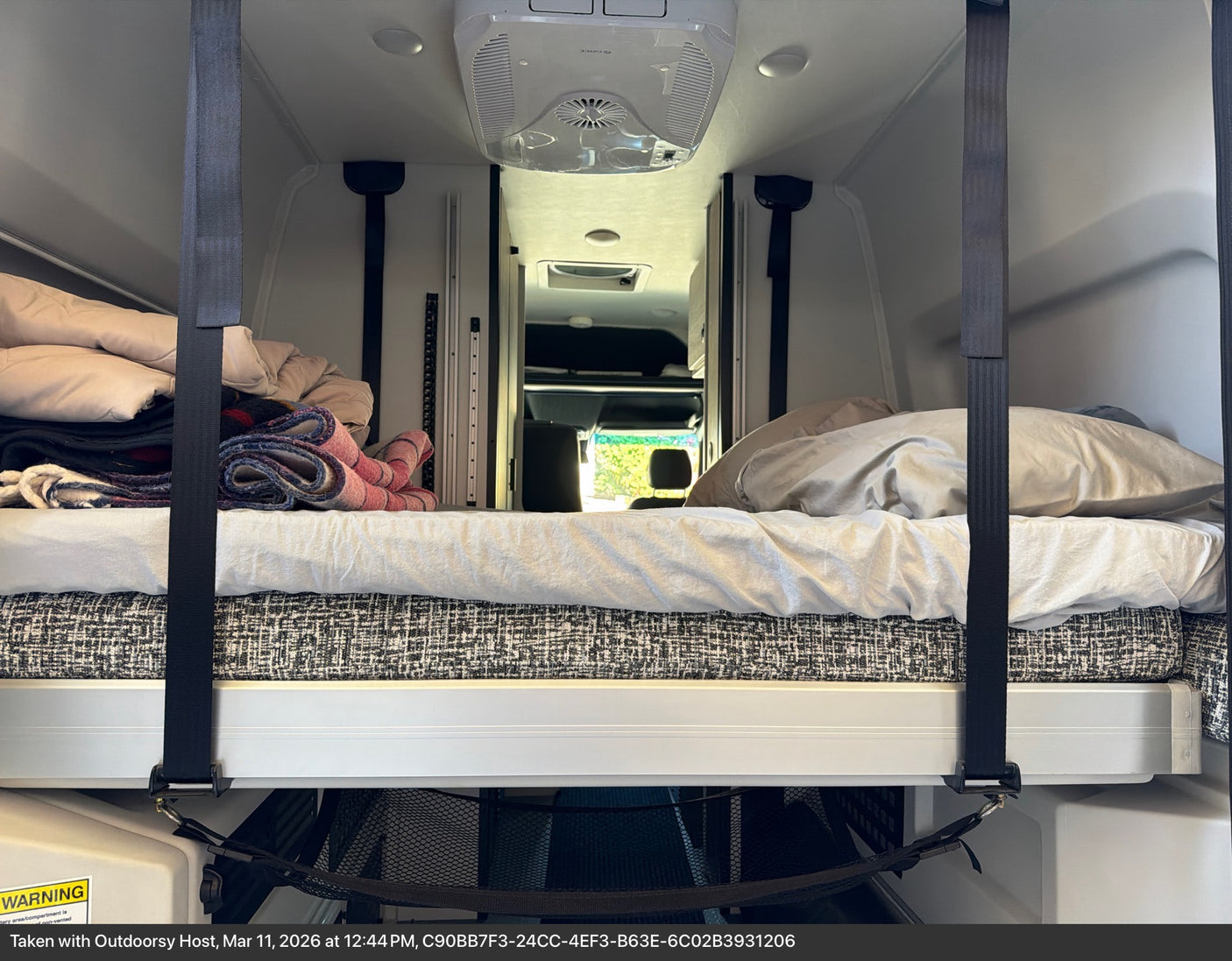 Cozy RV rental interior with a raised bed, pillows, blankets, and overhead AC. Straps secure the bed, storage net below.