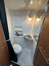 Compact RV rental bathroom featuring a toilet, corner sink, handheld shower, and shelving. A black shower curtain is tied back.