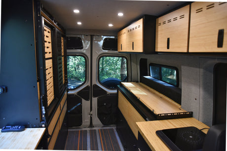 Inside this RV rental: a custom camper van with bamboo cabinets, built-in cooktop, and adaptable sleeping area, surrounded by forest views.