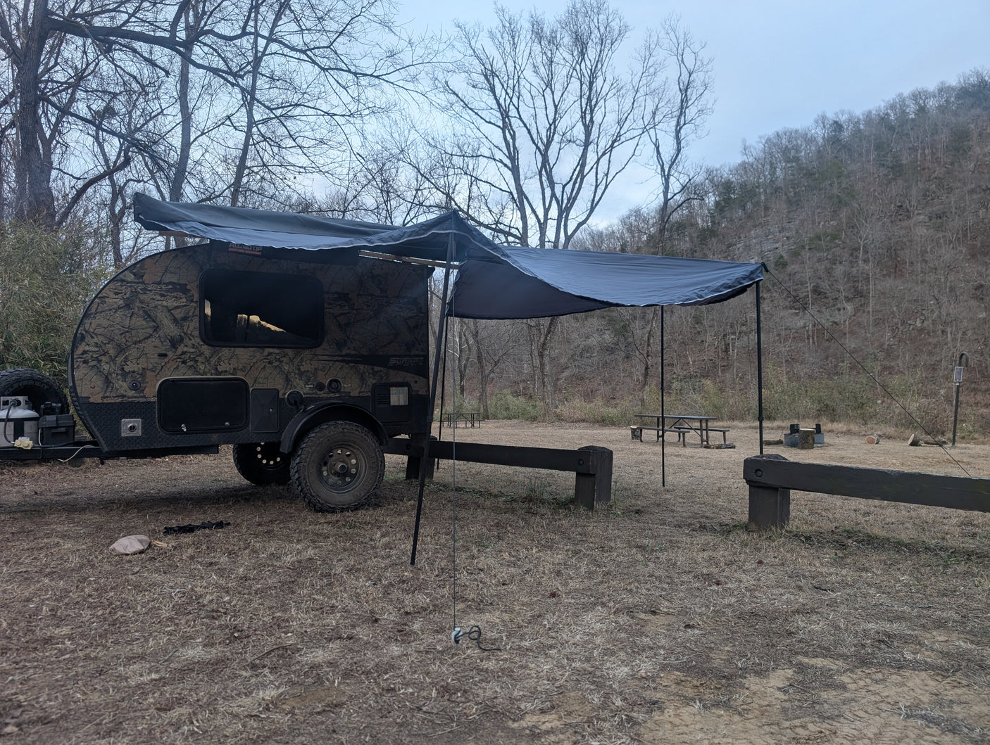 Adventure-ready RV rental: A camouflage teardrop camper with awning at a rustic campsite, surrounded by bare trees.