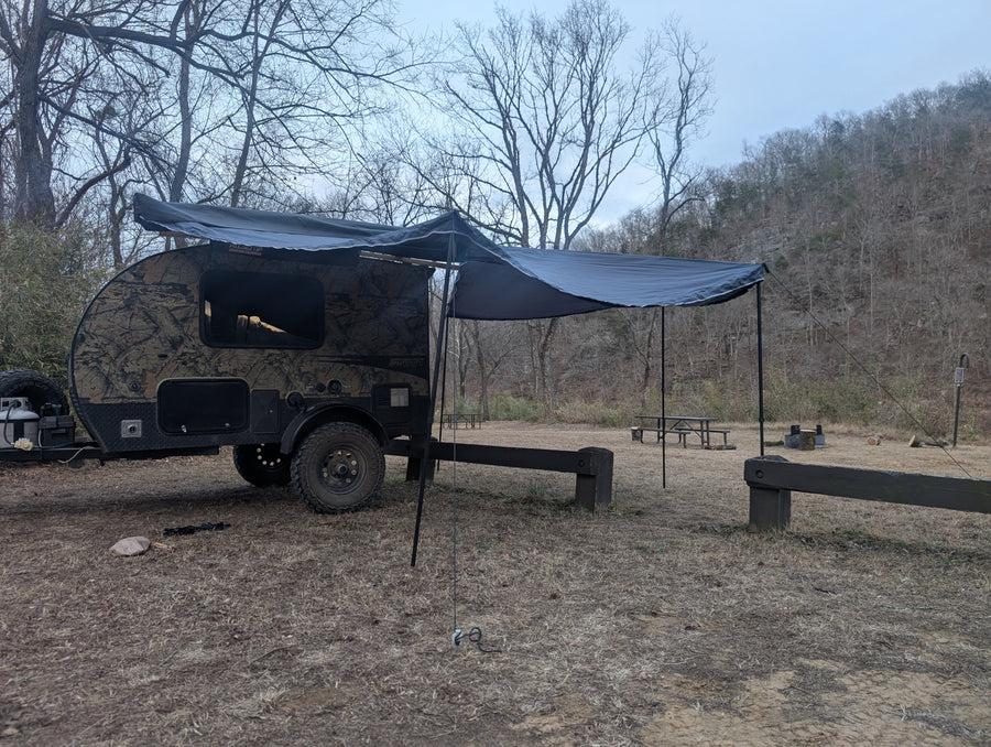 Adventure-ready RV rental: A camouflage teardrop camper with awning at a rustic campsite, surrounded by bare trees.