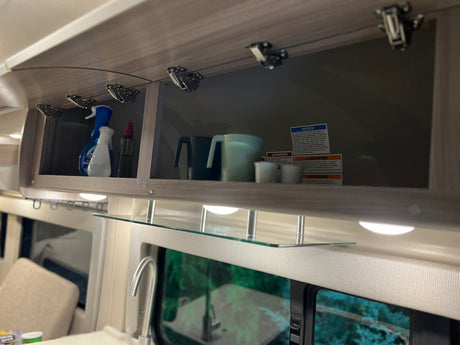 RV rental overhead cabinet organized with mugs, spray bottles, and safety warnings, plus a glass shelf below.