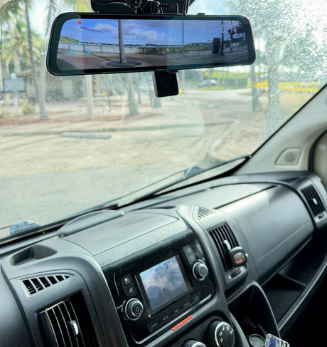 Dashboard and rain-splashed windshield of an RV rental. A digital rearview mirror shows a wide, scenic waterfront view.