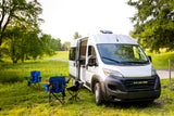 White RV rental camper van with open door, camping chairs, and a table in a grassy outdoor setting.