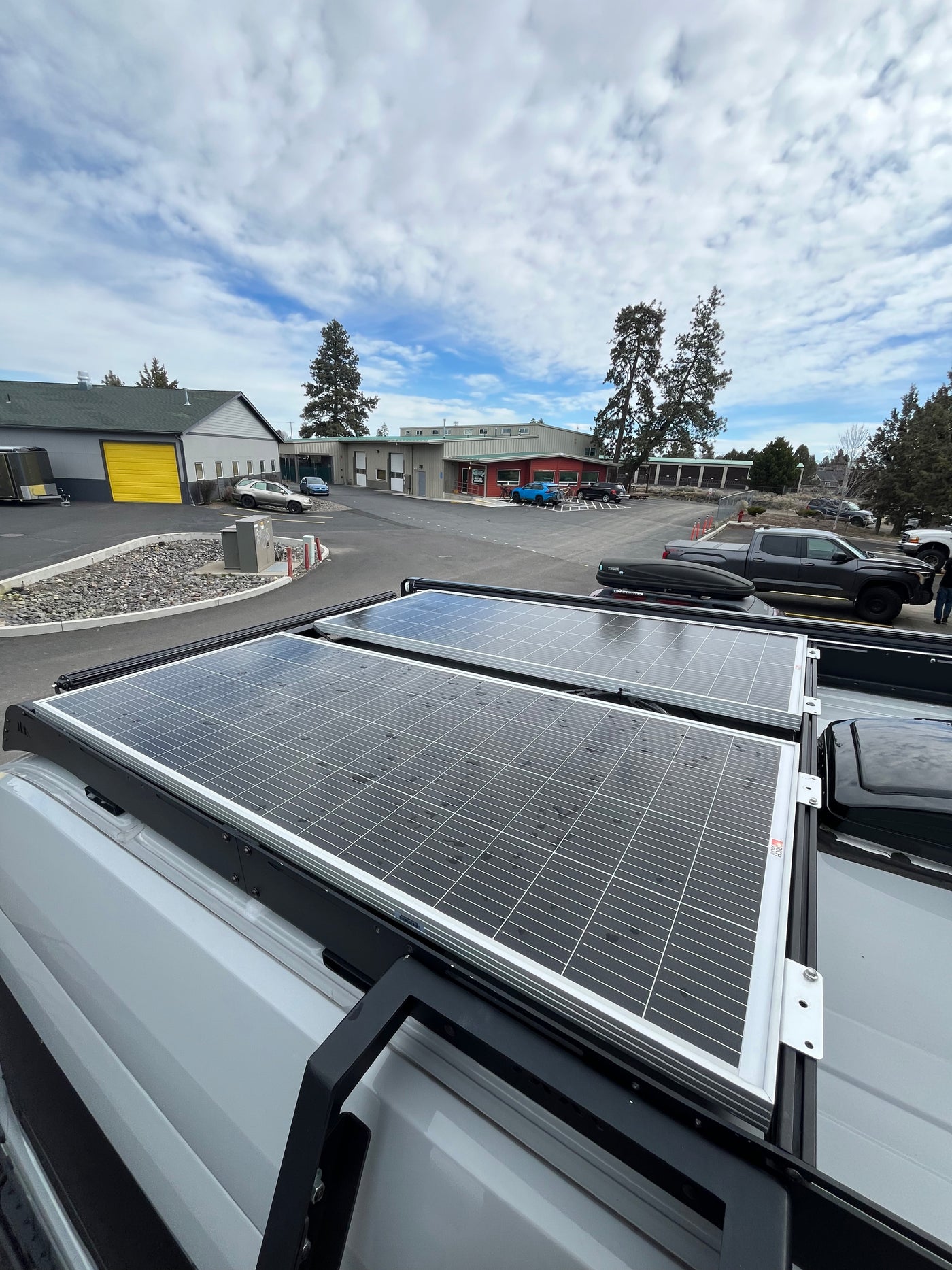 Two large solar panels on an RV rental van rooftop, with commercial buildings & parked cars in the background under cloudy skies. — photo 6