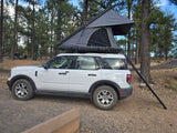 White SUV with a rooftop tent deployed in a wooded campsite, offering a unique RV rental experience.