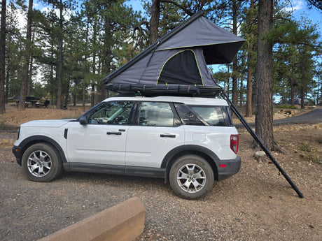 White SUV with a rooftop tent deployed in a wooded campsite, offering a unique RV rental experience.