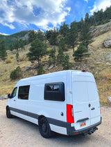 White Mercedes-Benz Sprinter camper van parked on a dirt road in a treed mountain landscape, great for an RV rental.