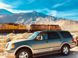 A gray SUV parked near train tracks, with a yellow train, wind turbines, and mountains under a blue sky, perfect for an rv rental adventure.