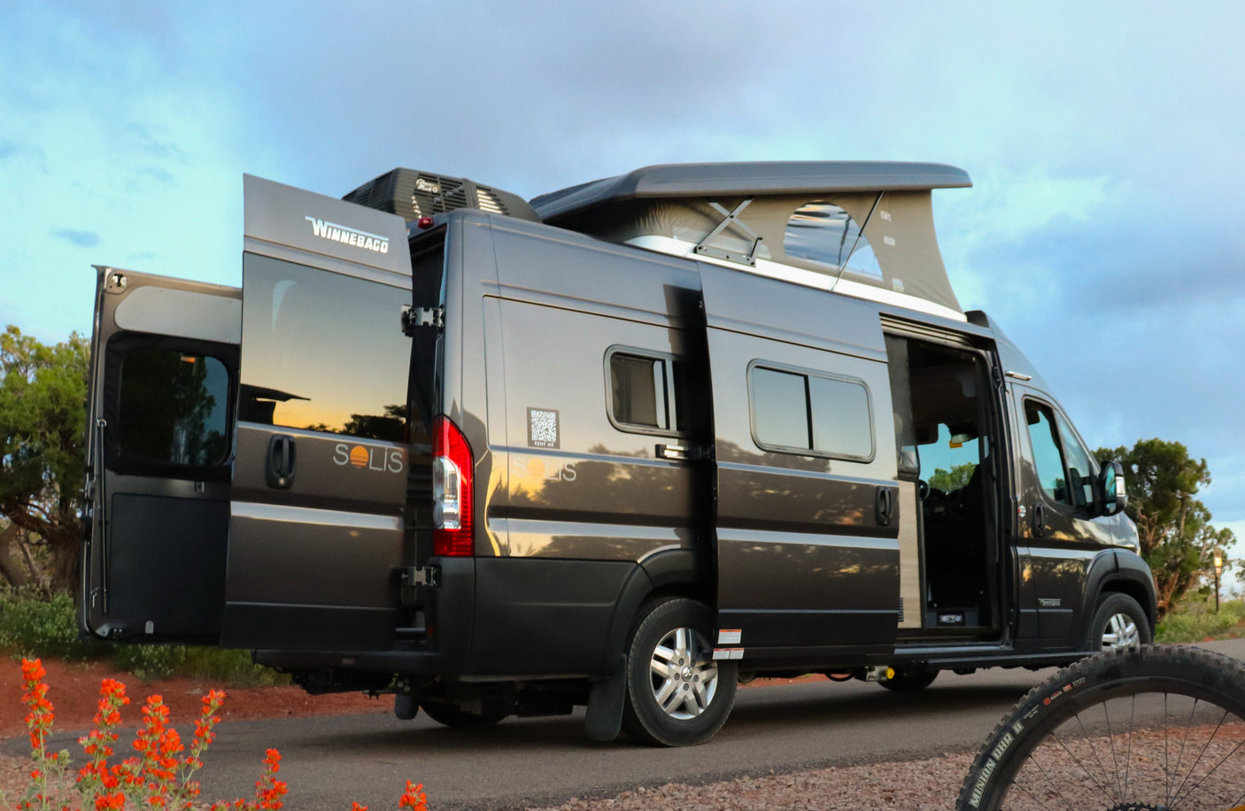 A gray Winnebago Solis camper van, great for RV rental and van life adventures, with open doors & pop-top extended at sunset.