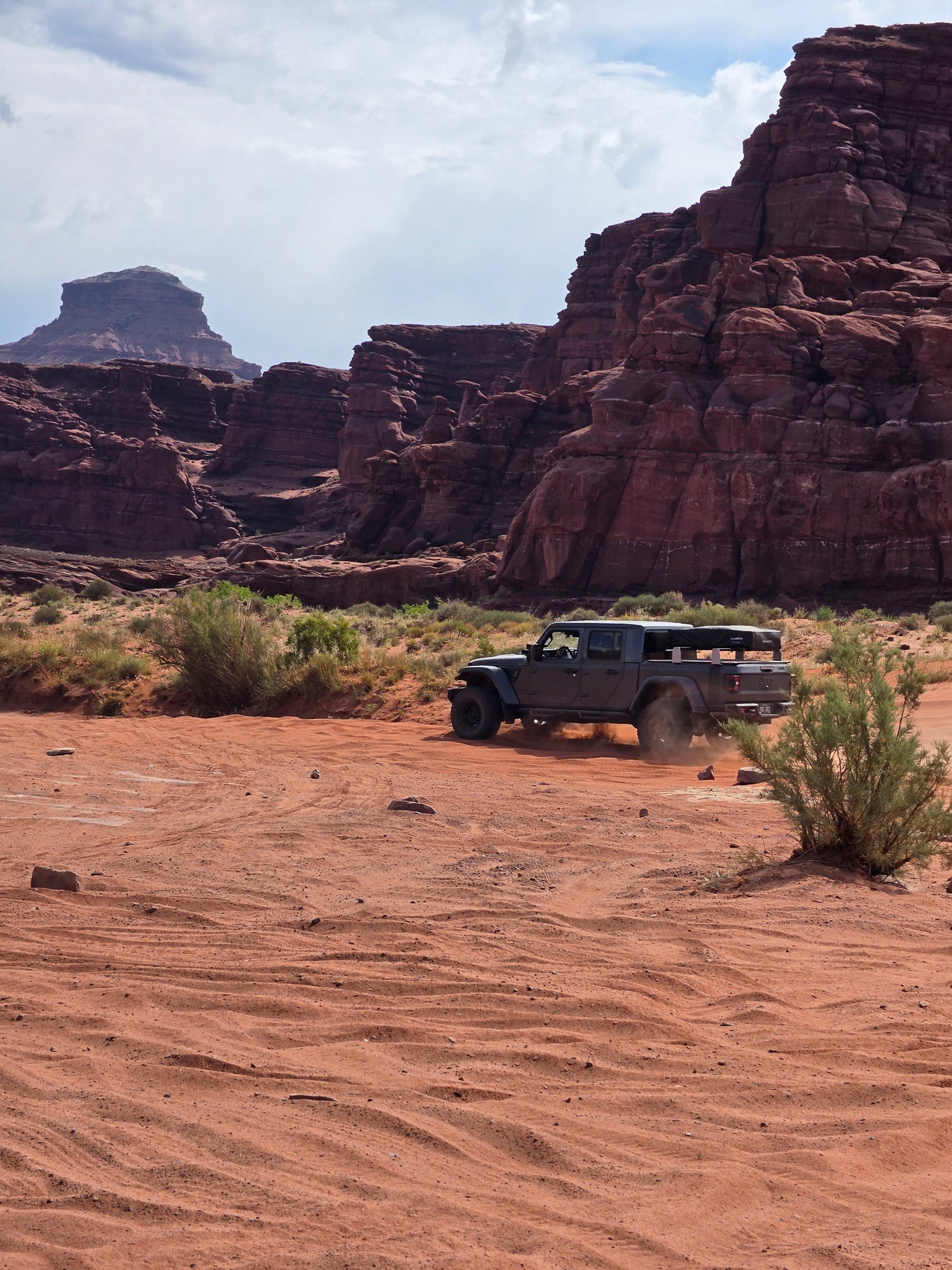 Jeep Gladiator with rooftop tent kicks up dust on a desert trail among red rock formations. Explore with an RV rental adventure!