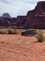Jeep Gladiator with rooftop tent kicks up dust on a desert trail among red rock formations. Explore with an RV rental adventure!
