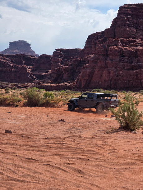 Jeep Gladiator with rooftop tent kicks up dust on a desert trail among red rock formations. Explore with an RV rental adventure!