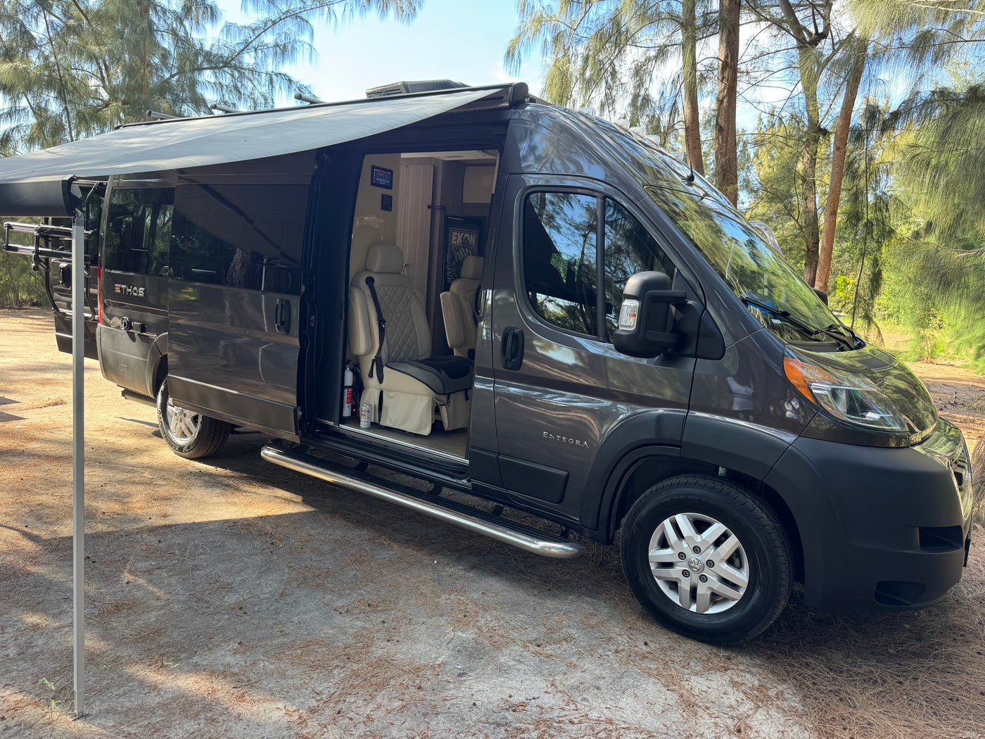 Dark gray Entegra Ethos camper van rv rental with an extended awning, parked in a pine forest. The side door is open, showing the interior. — photo 17