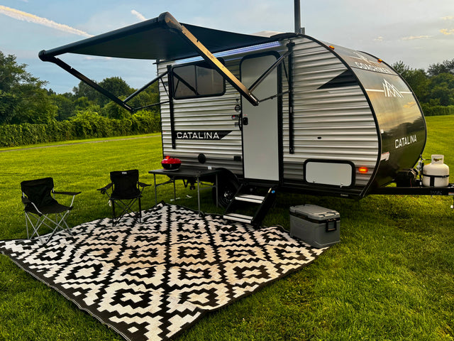 Experience the outdoors! An rv rental travel trailer with an extended awning, chairs, grill, and patterned rug on green grass.