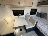 Stylish RV rental interior: dinette with marbled table, plush benches, and bed with patterned bedding, plus overhead cabinets.