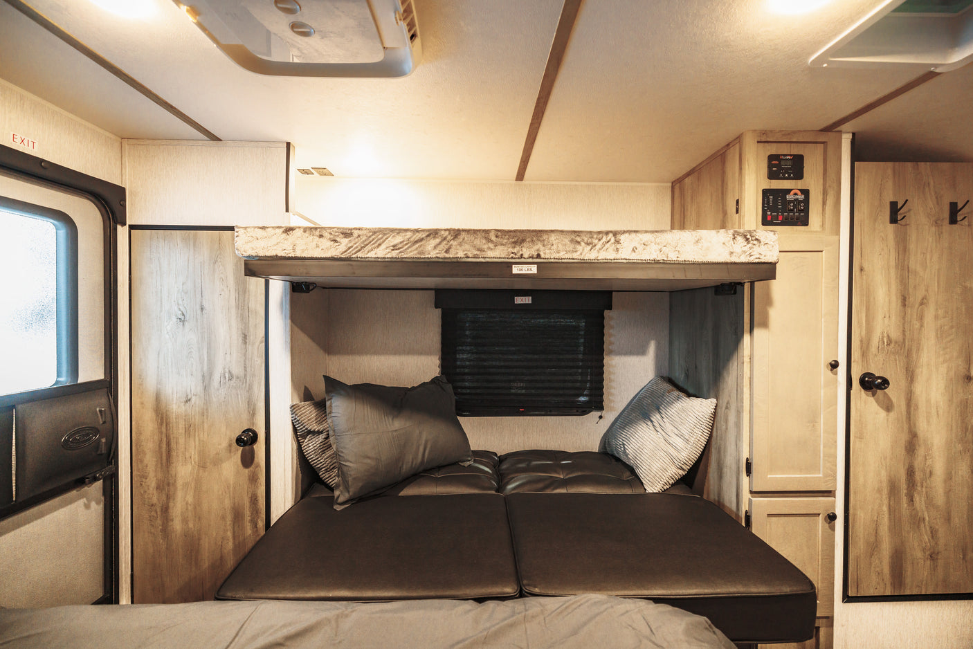 Inside this cozy RV rental: lower bunk with seating, upper bunk (100 lbs cap), exit, and essential control panels.