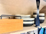 Organized RV rental kitchen with stacked plates & bowls in white, gray, and black, a wooden caddy, and metal shelf divider.