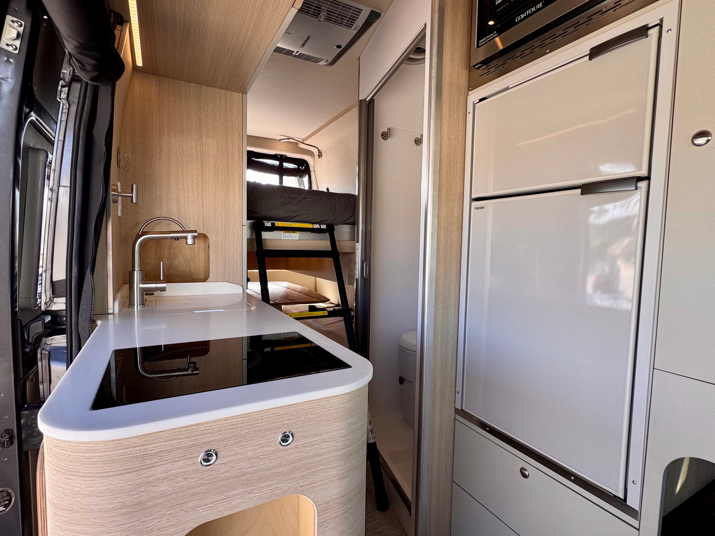 Compact rv rental interior with kitchenette (sink, cooktop), refrigerator, microwave, and bunk beds, plus a small bathroom.