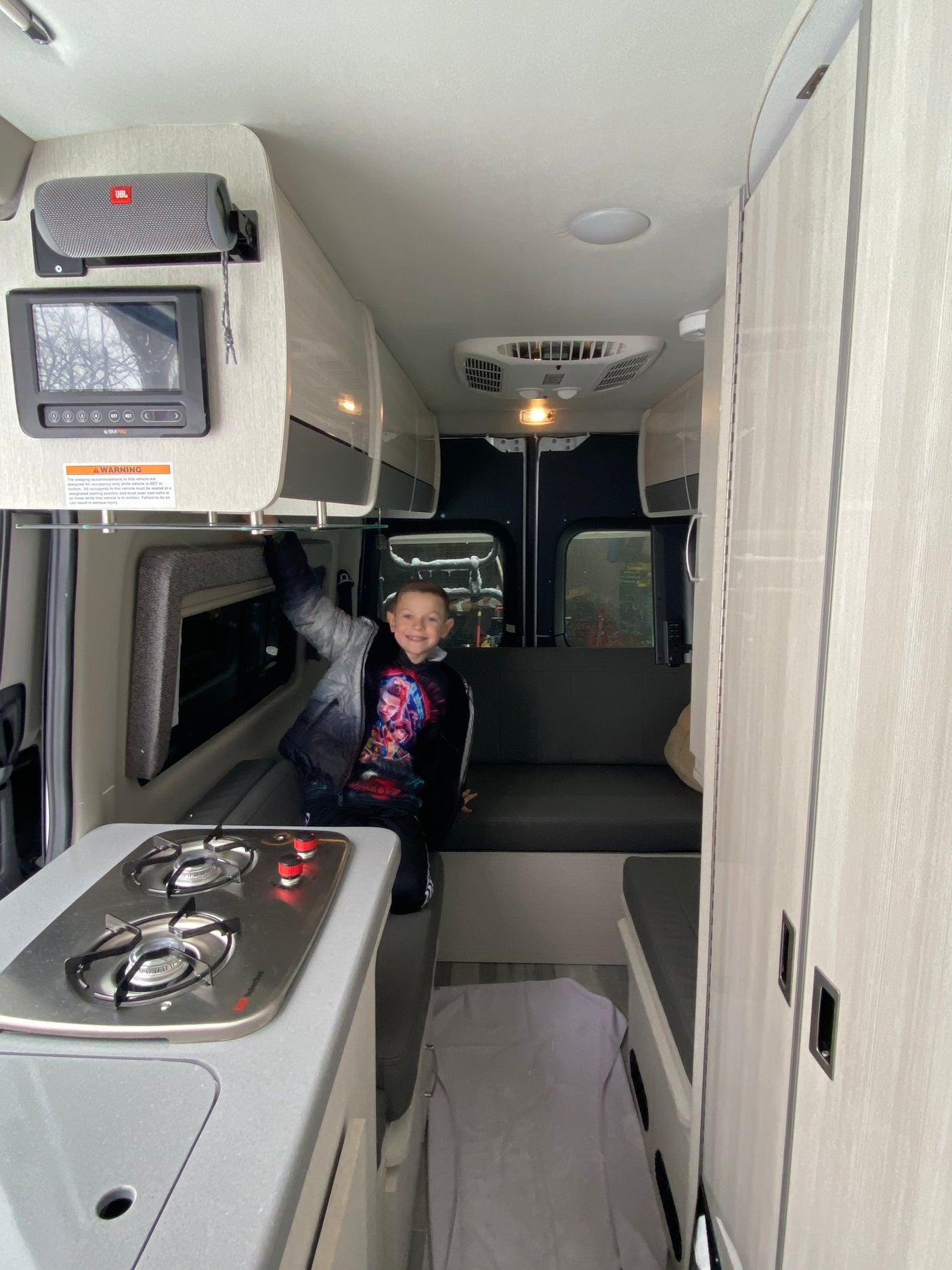 A cheerful boy poses inside a modern RV rental. View shows compact kitchen with stovetop, seating, and overhead storage.