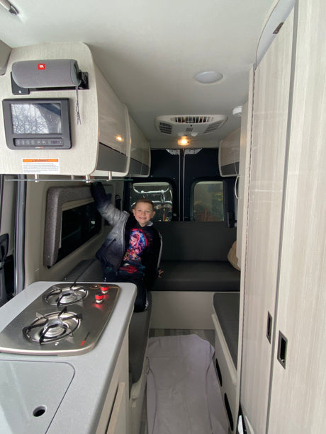 A cheerful boy poses inside a modern RV rental. View shows compact kitchen with stovetop, seating, and overhead storage.