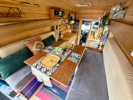 Cozy RV rental's wooden interior, featuring a dining table set for two, cushions, a kitchenette, and forward view.