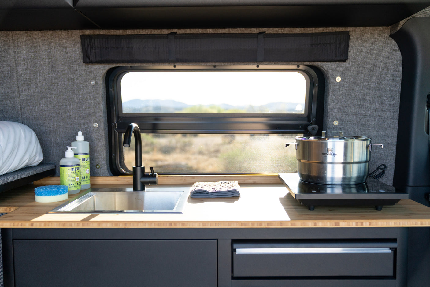 An RV rental's functional kitchen area: sink, black faucet, induction cooktop, and window views for your travels.