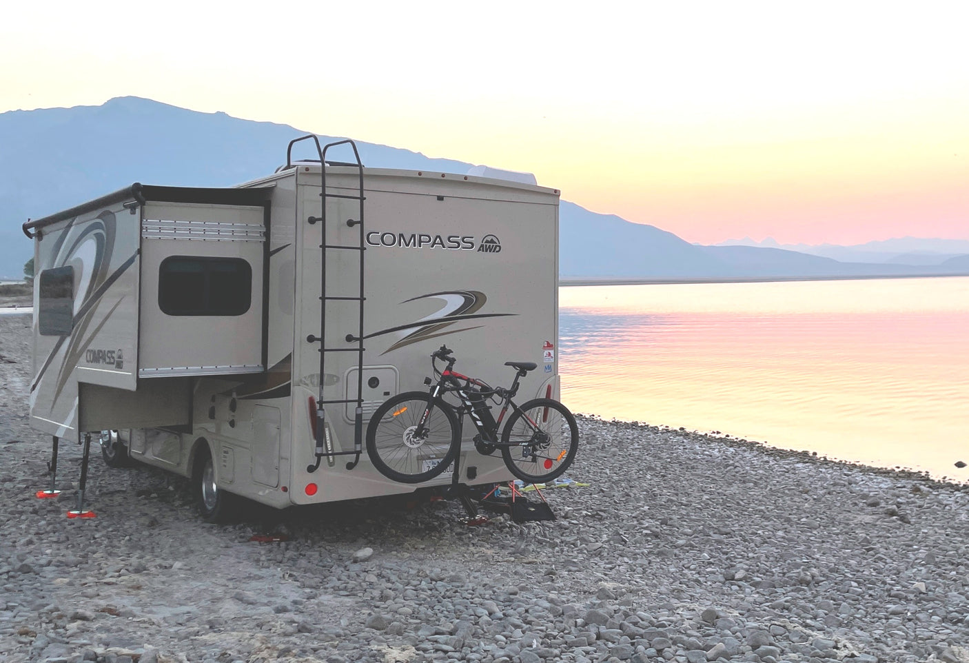 An RV rental, complete with bike, parked lakeside at sunset, showcasing serene nature and travel possibilities.